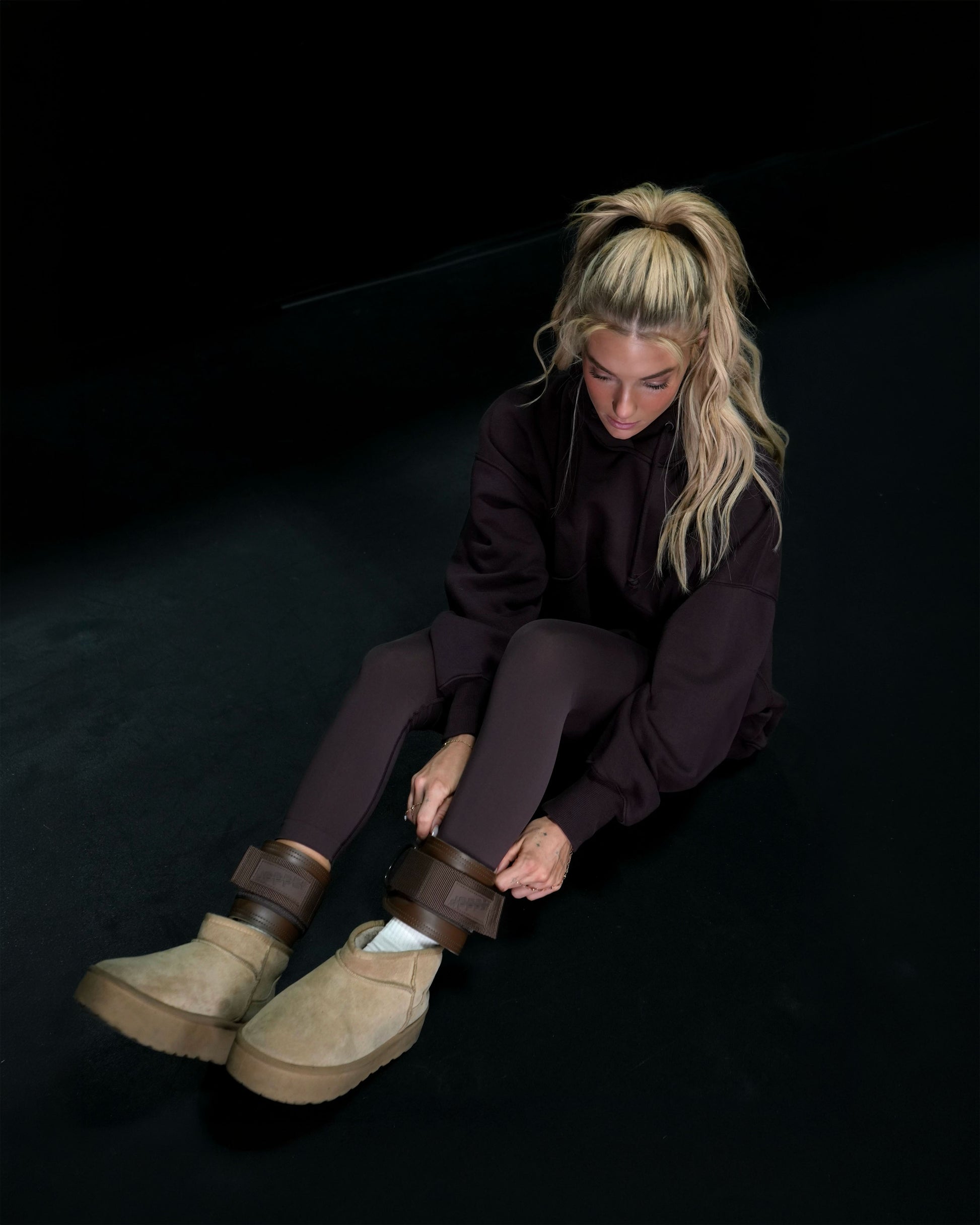 athlete sitting on dark floor strapping on umber uppper ankle straps 2.0 fitness gear