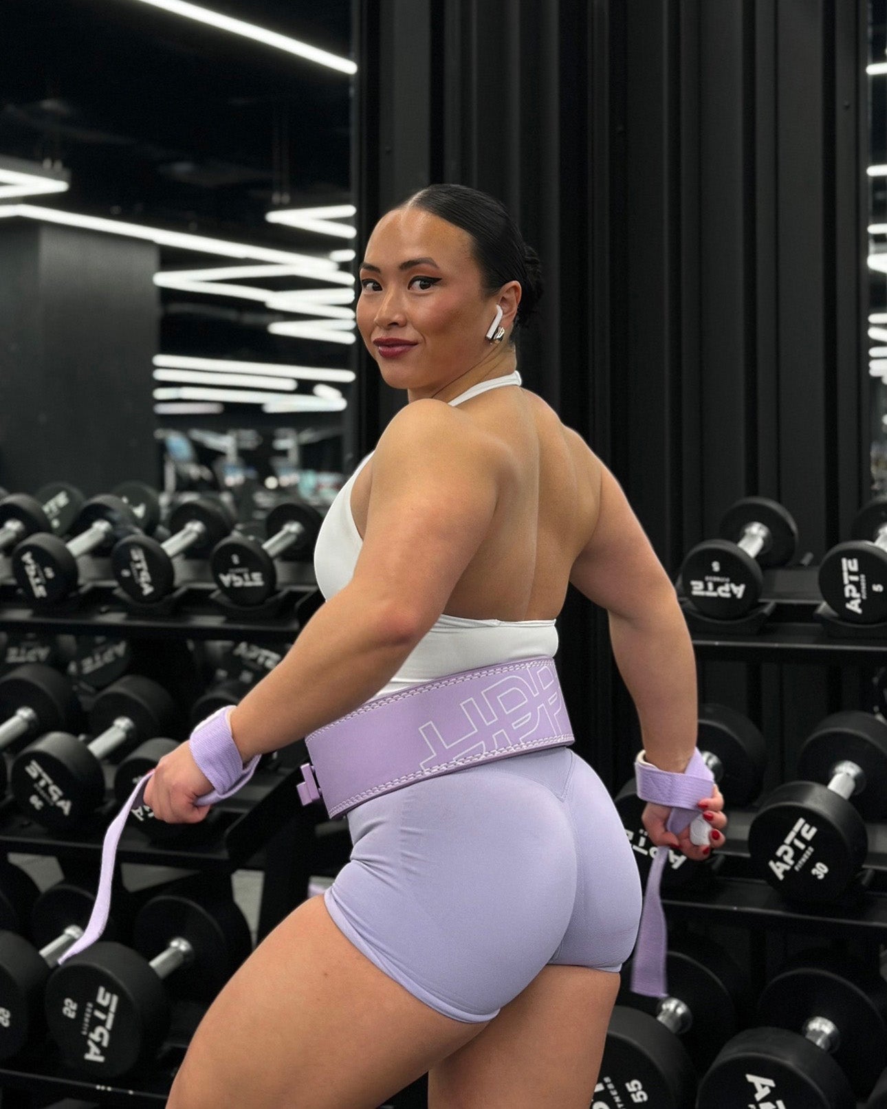 athlete in lavender gym outfit posing from behind wearing lavender uppper lever belt near dumbbell rack in gym