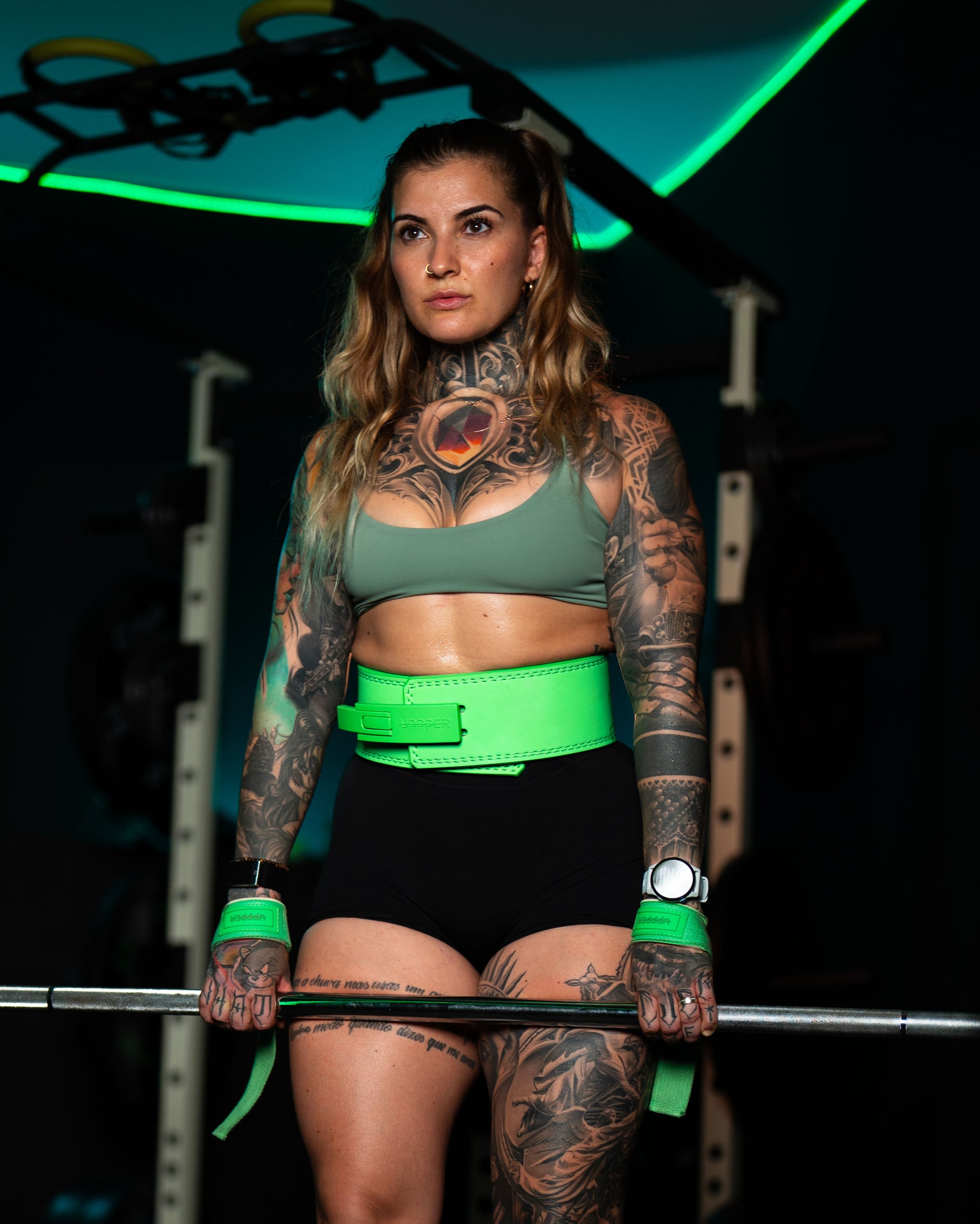 tattooed athlete wearing neon green uppper lever belt and wrist wraps at barbell in dark gym