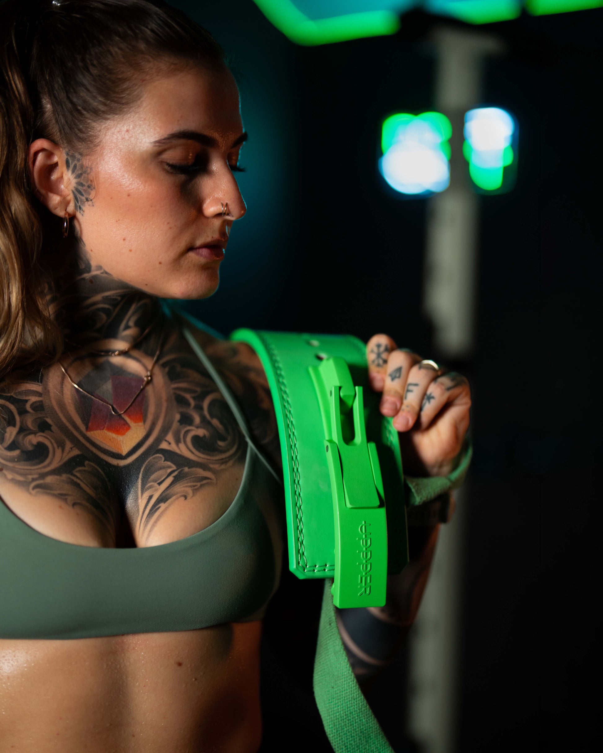 tattooed athlete in green sports bra looking at neon green uppper lever belt against dark background