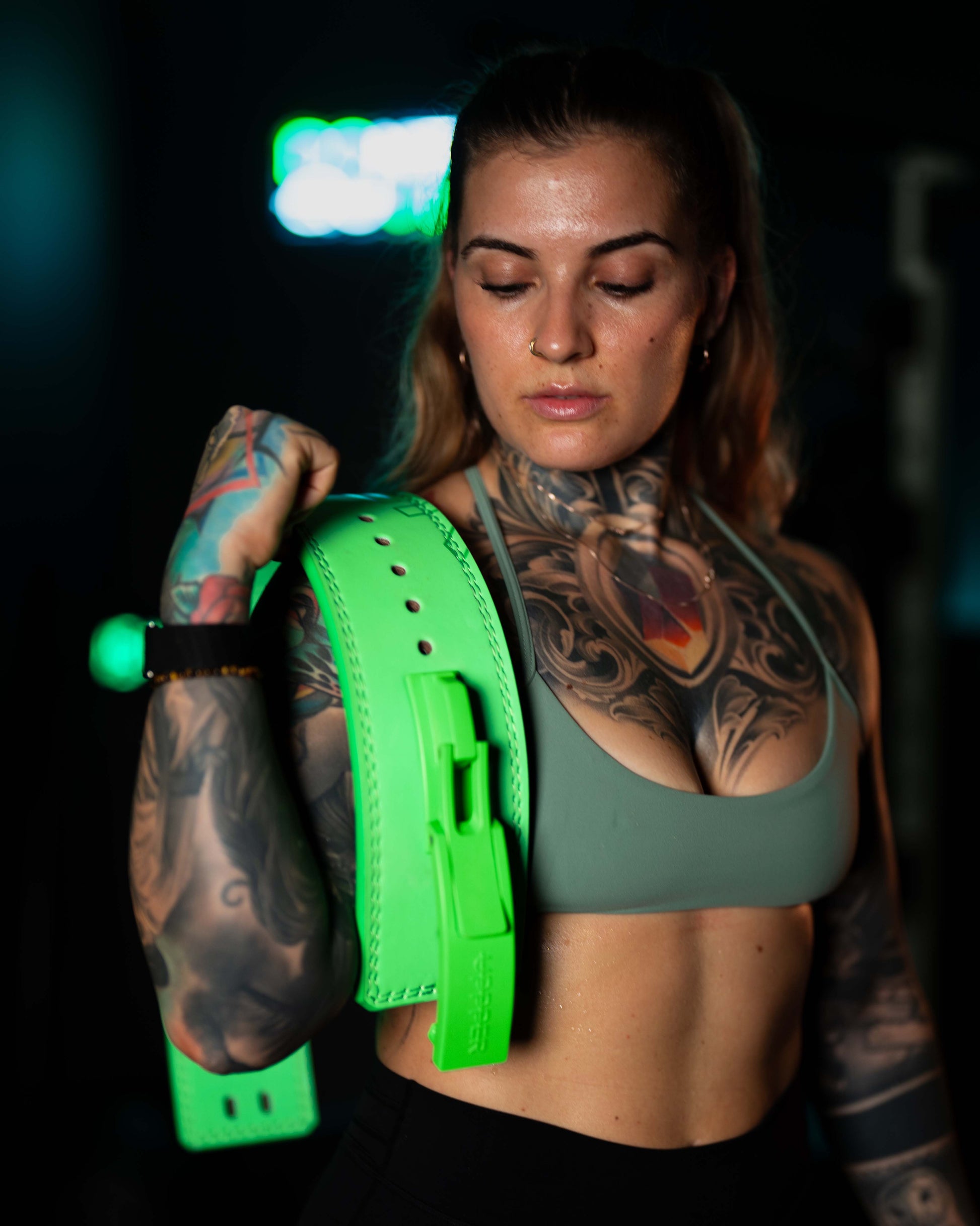 tattooed athlete in green sports bra holding neon green uppper lever belt over shoulder against dark background