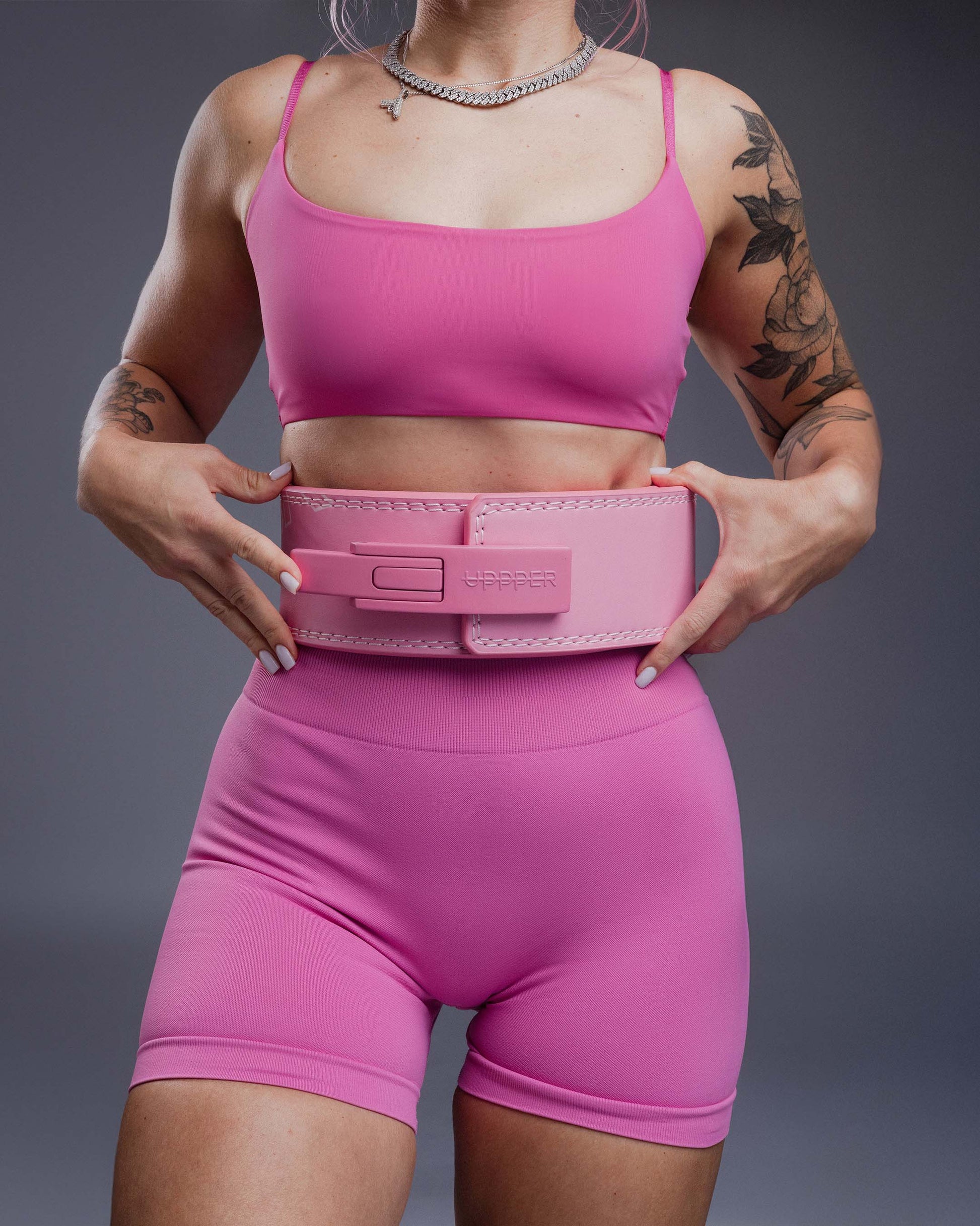 tattooed athlete in pink gym outfit posing with pink uppper lever belt on gray background