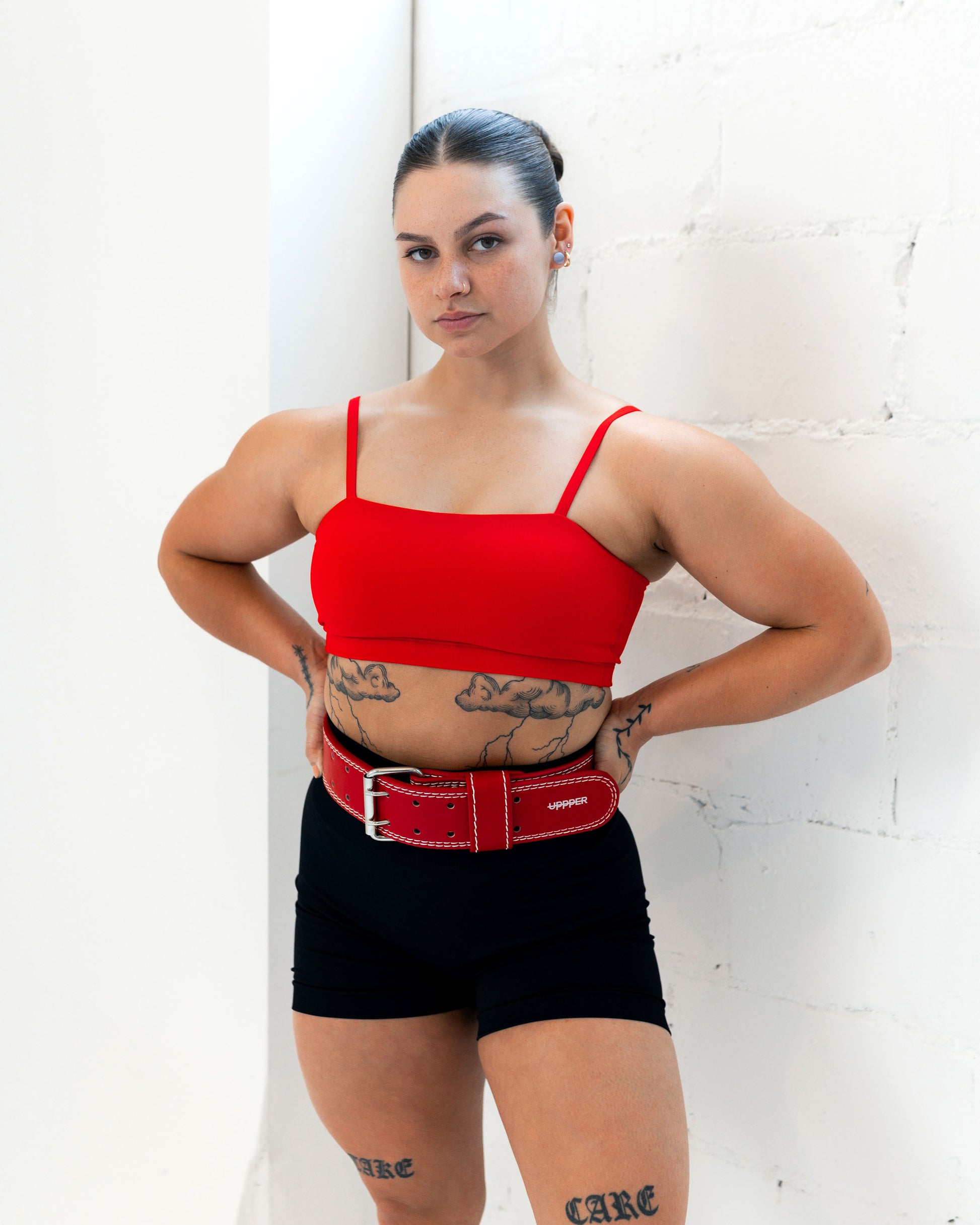 woman wearing a red crop top and black shorts with a red uppper lifting belt against a white wall